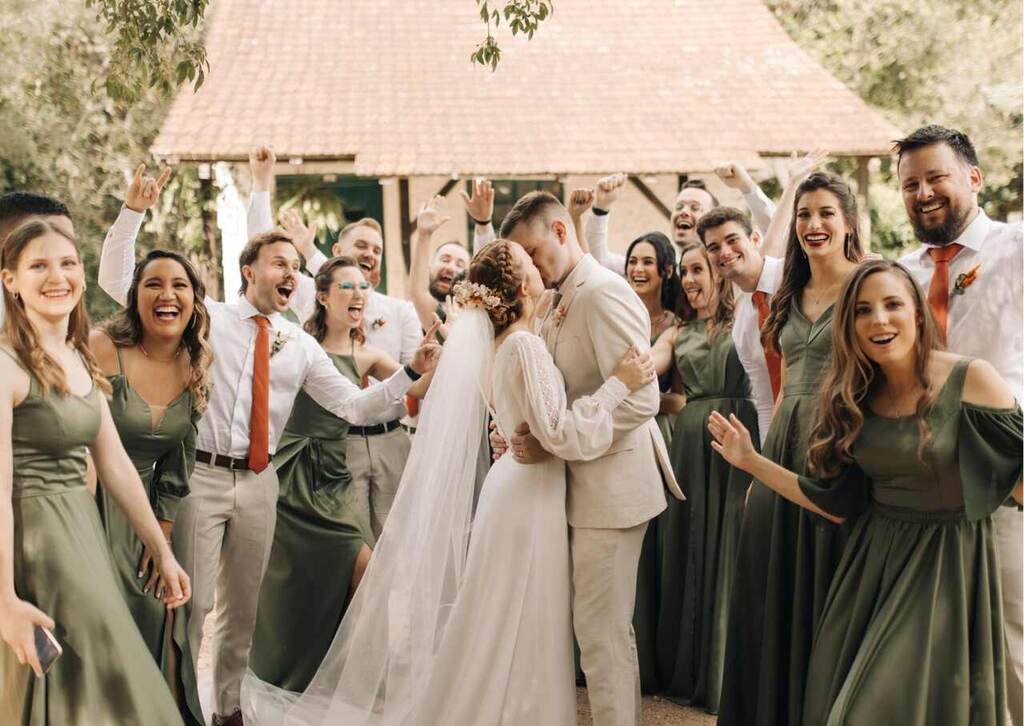 noivos se beijando enquanto madrinhas com vestido verde e padrinhos com calça bege camisa branca e gravata laranja comemoram