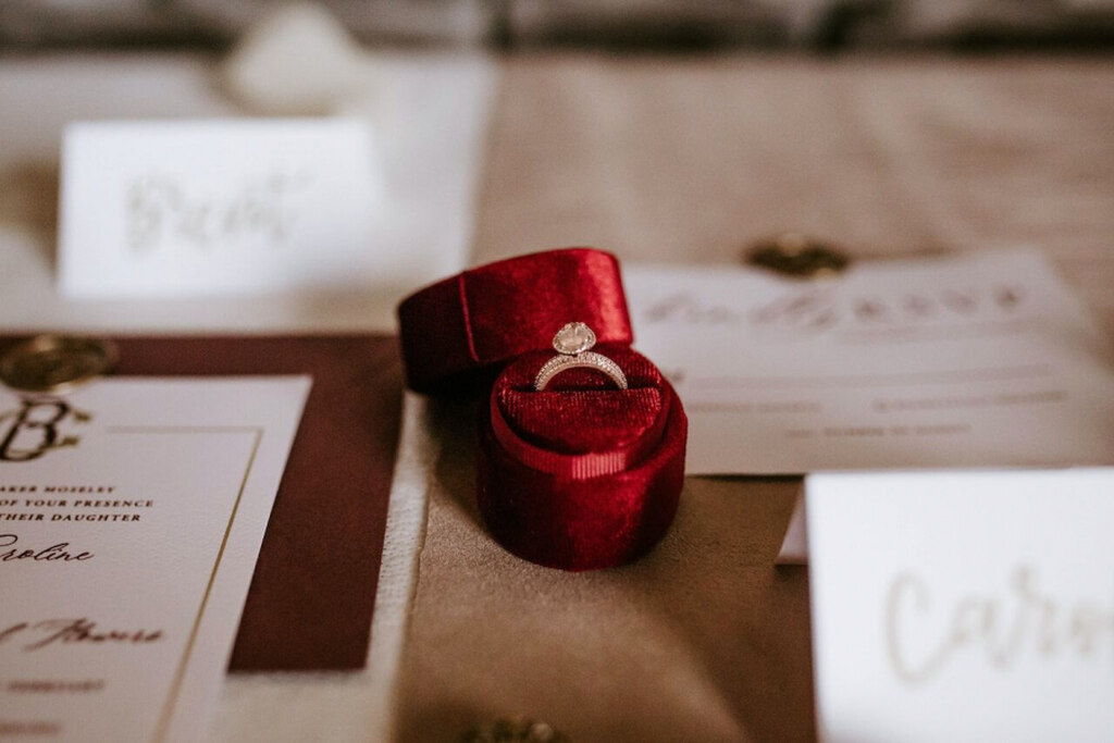 caixa vermelha de veludo com aliança com pedra brilhante e papelaria de casamento ao lado em cima da mesa