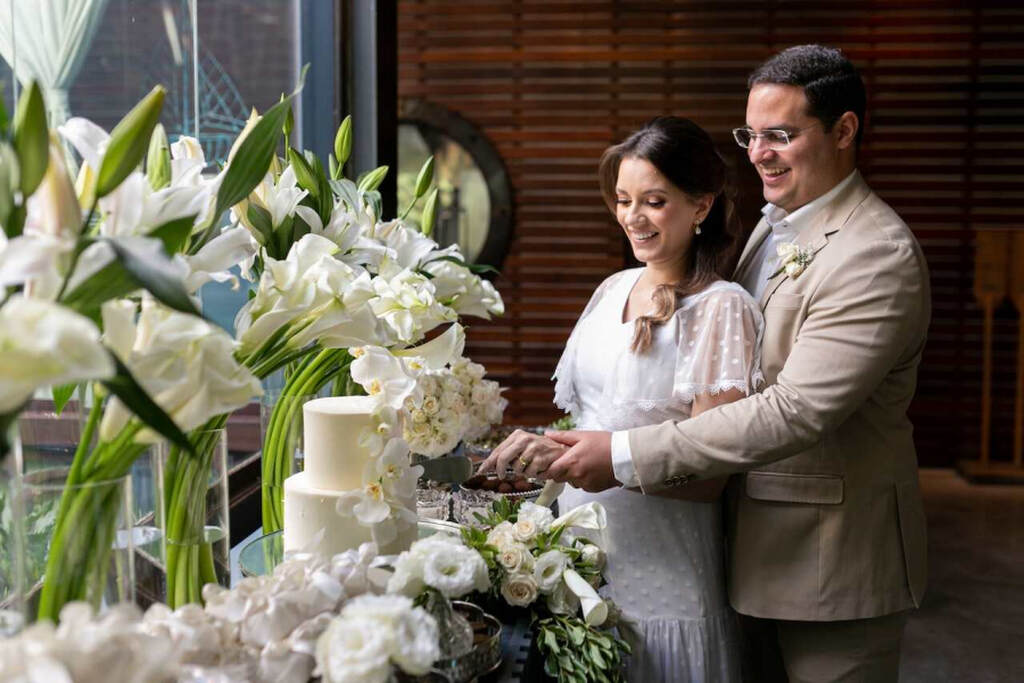 noivos cortando o bolo de casamento branco em cima da mesa com vasos com flores de copo de leite e rosas 
