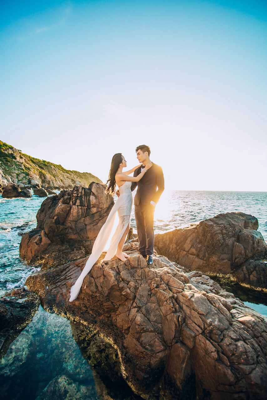 mulher comemorando bodas de casamento com vestido branco em cima de pedra a beira do mar com o marido