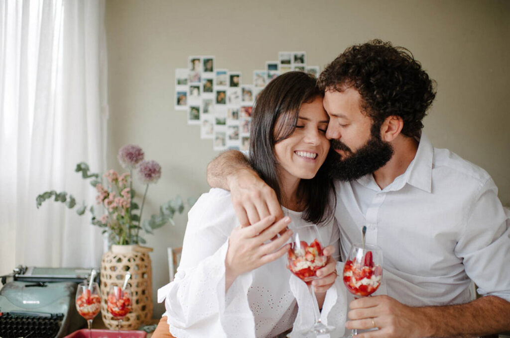 casal abraçado dentro de casa segurando taça com morango e chantilly