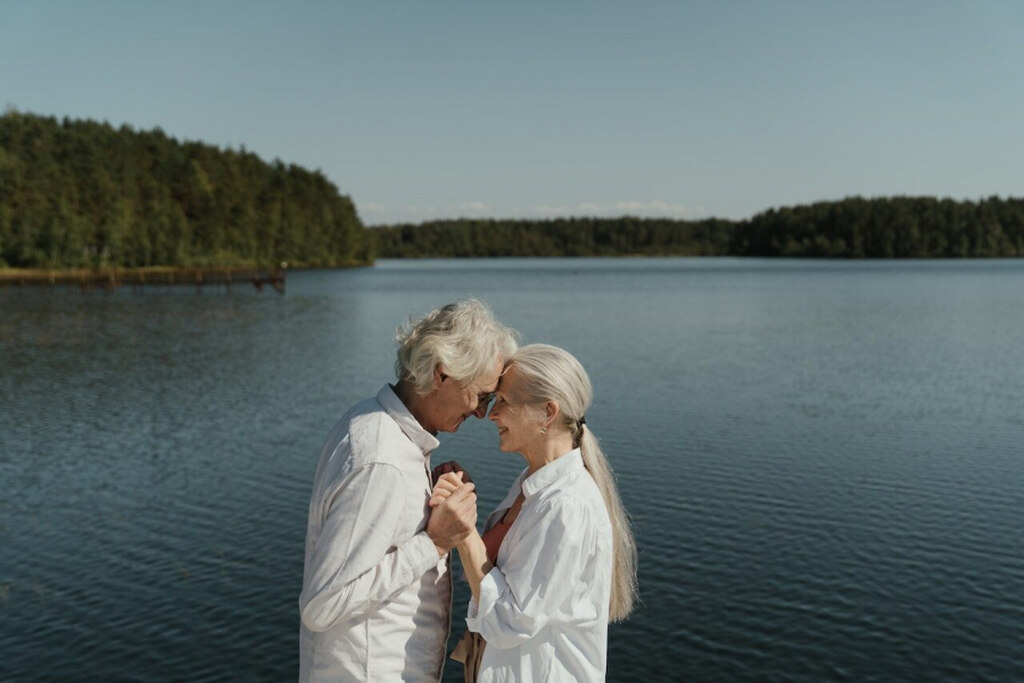 casal idosos com camisas brancas de mãos dadas na frente do rio