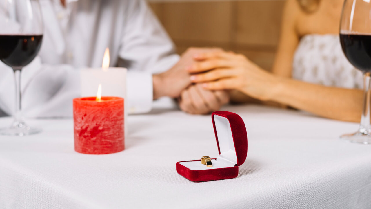 casal de mãos dadas em cima da mesa com taça de vinho vela vermelha e porta-aliança vermelho