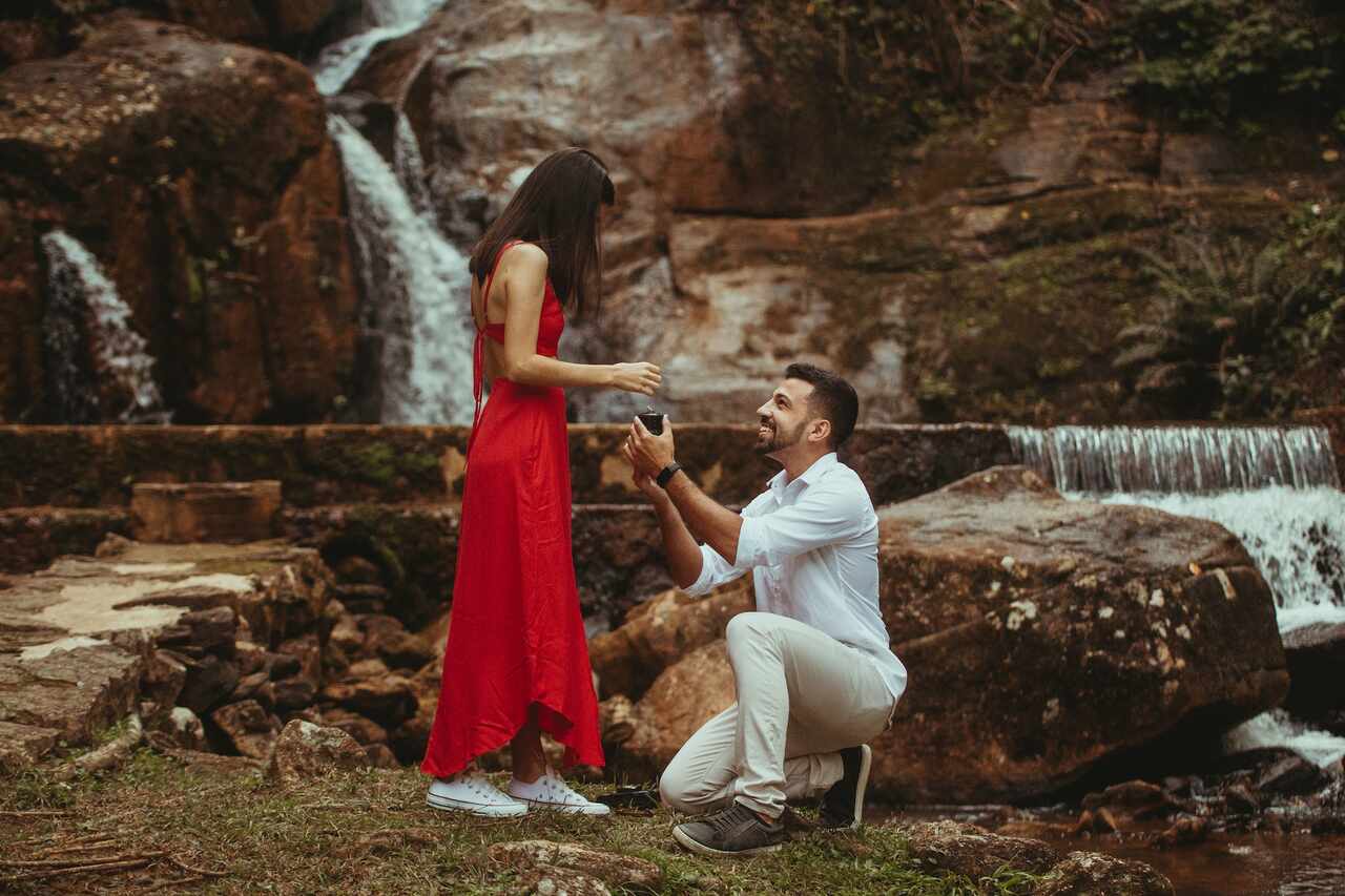 homem com roupa social clara ajoelhada perto de cachoeira fazendo o pedido de casamento para noiva com vestido vermelho e all star branco