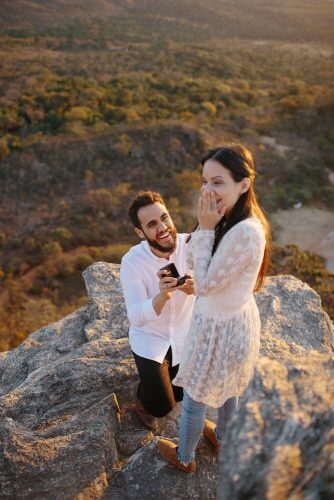 Homem com camisa branca sorrindo no topo de uma pedra fazendo o pedido de casamento