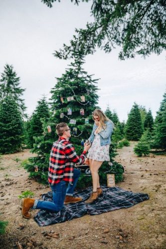 homem com camisa xadrez fazendo pedido de casamento em frente a pinheiro com decoração de natal e mulher com vestido branco com casaco jeans