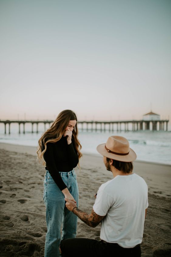 homem de joelhos na praia com camiseta branca e chapéu fazendo pedido de casamneto para mulher com calça jeans e blusa preta na praia
