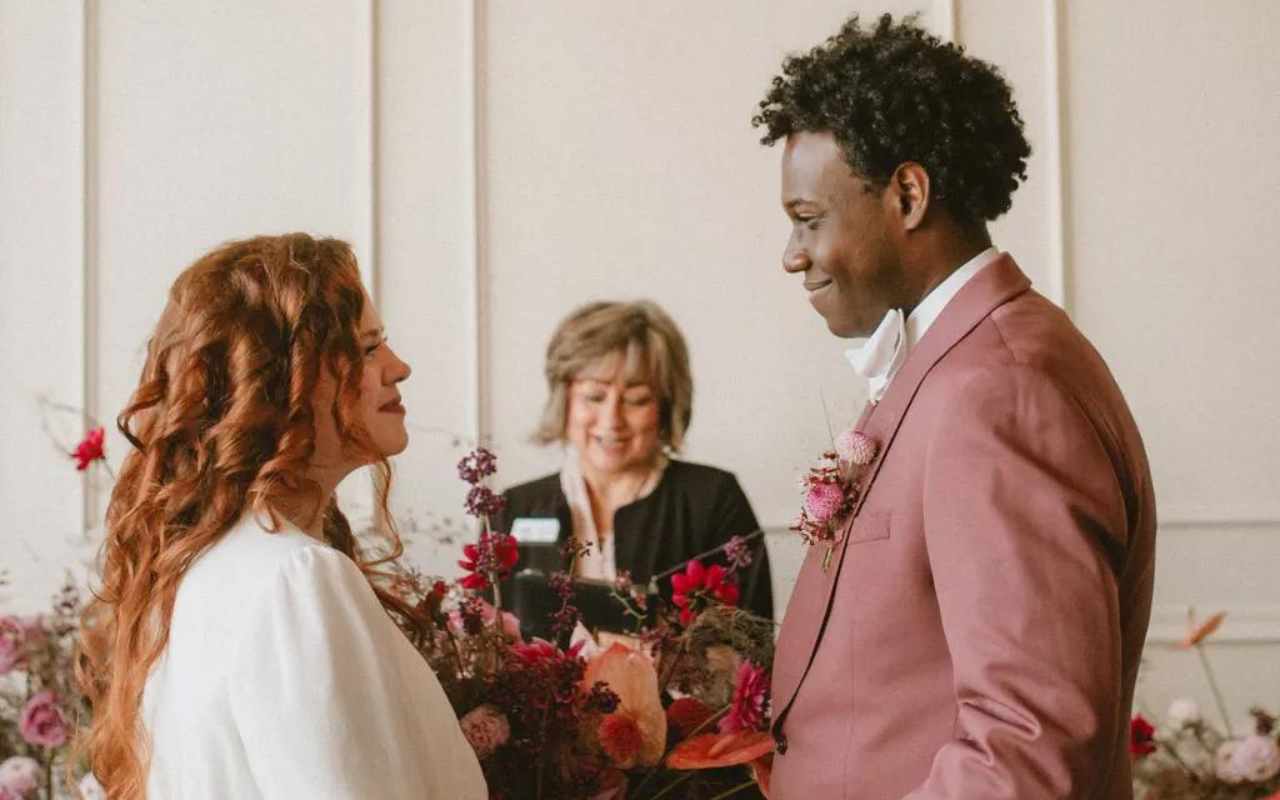 noiva ruiva com noivo negro em frente ao altar de casamento durante cerimônia de casamento conduzida por celebrante
