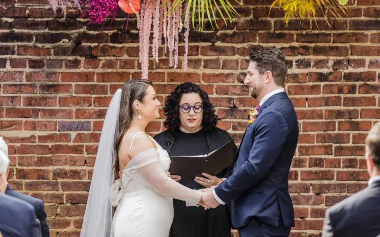 casal de noivos de mãos dados em altar de casamento debaixo de arranjo floral colorido ouvindo as palavras do celebrante de casamento
