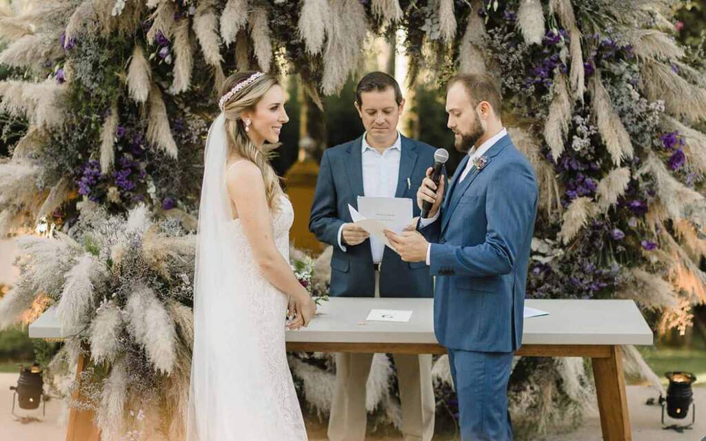 noivo falando votos de casamento olhando para noiva vestida de branco em altar com capim dos pampas e flores roxas. Atrás do aparador 