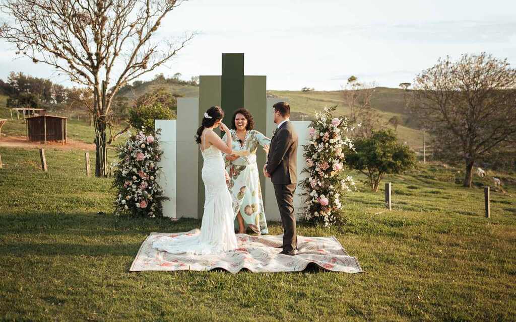 cerimônia de casamento ao ar livre no gramado de uma fazenda. Os noivos juntamente com a celebrante de casamento estão em frente a um altar moderno em tons de verde e flores rosas