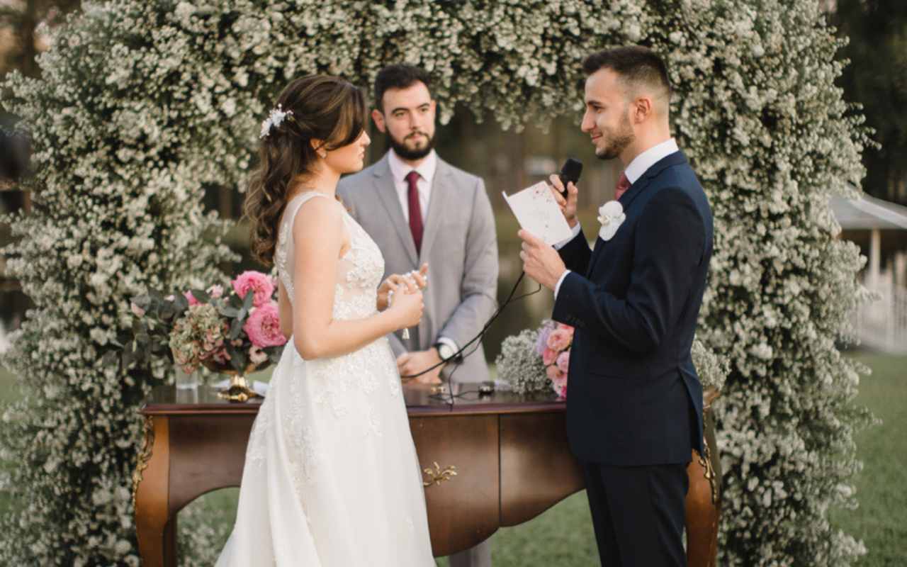 noivo dizendo votos de casamento em frente a um altar com flores brancas. atrás do aparador de madeira, está o celebrante de casamento
