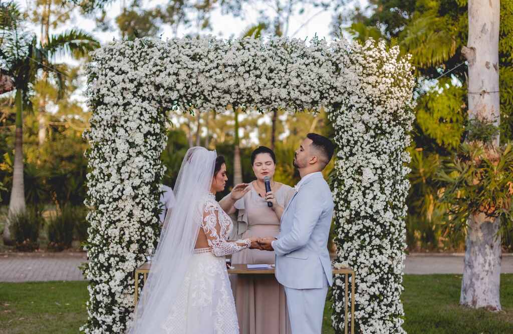 casal de noivos em frente a um altar quadrado com flores de mosquitinho branca. Atrás encontra-se o celebrante de casamento