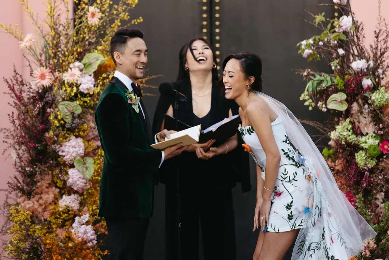 casal de noivos durante a troca de votos de casamento rindo juntamente com celebrante de casamento. o noivo veste terno de veludo verde e a noiva um vestido de noiva curto com bordados coloridos de flores