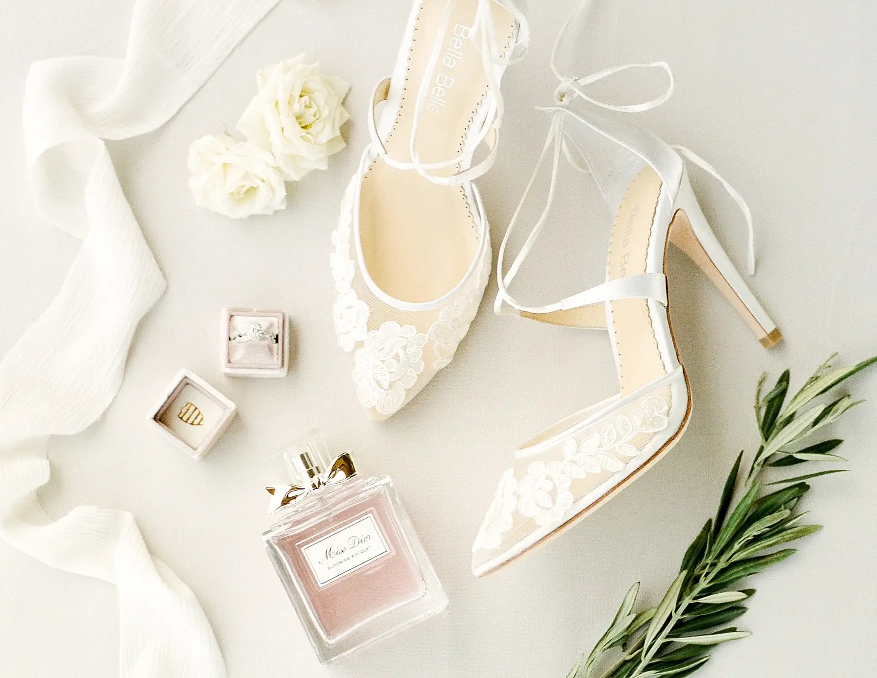 foto de sapato de noiva rendado branco de bico fino ao lado de alianças e perfume da noiva