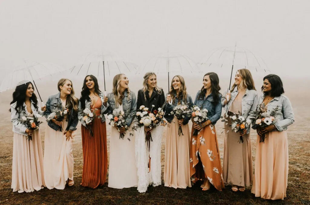 casamento na praia com chuva e madrinhas com guarda chuva transparente ao lado da noiva. Todas estão usando jaquetas jeans