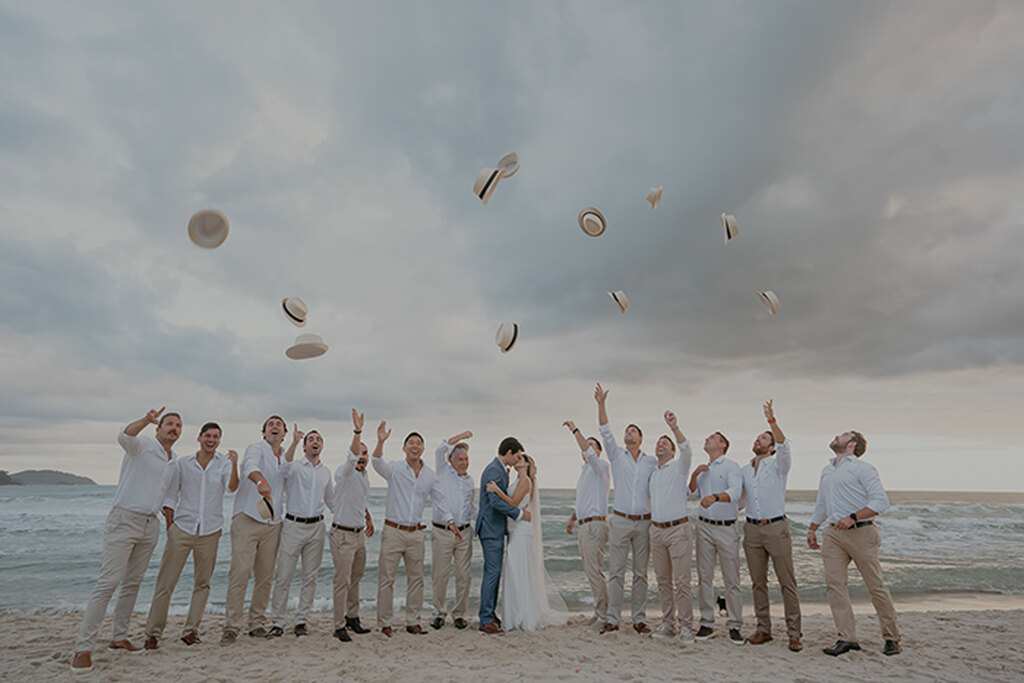 noivos se beijando em casamento na praia e em volta estão os padrinhos jogando os chapéus 