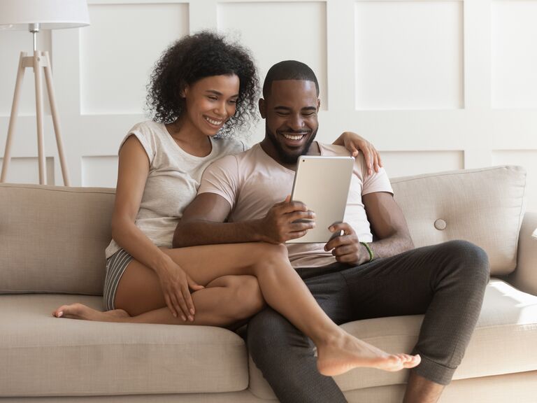 casal negro sorrindo sentado em sofá bege pesquisando a lista de casamento o que pedir