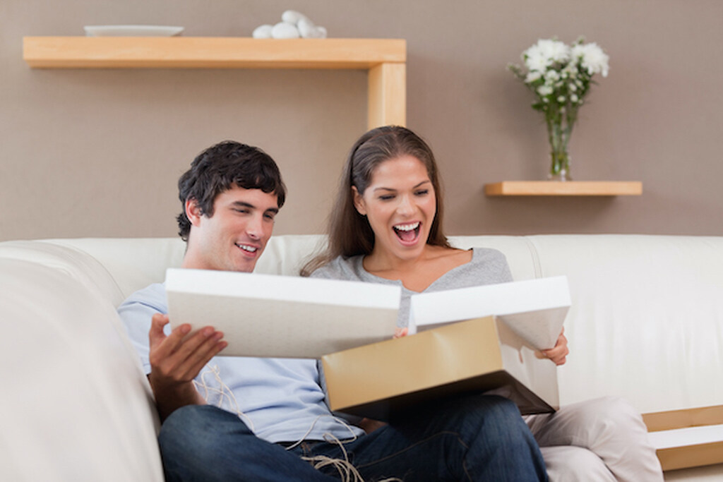 casal sentado no sofá branco sorrindo ao abrir caixa com presente da lista de casamento simples