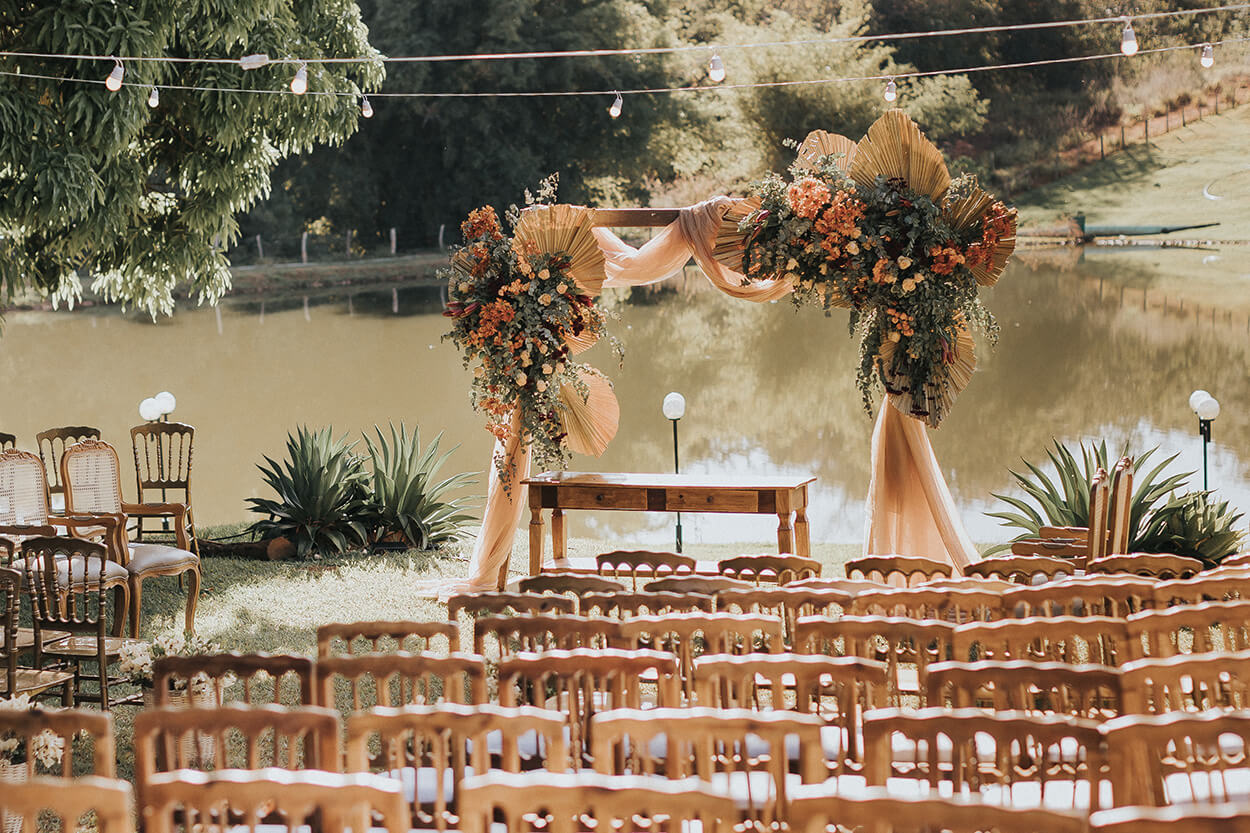 Cadeiras, mesa de madeira e arco com folhagem seca e flores laranja na beira do rio