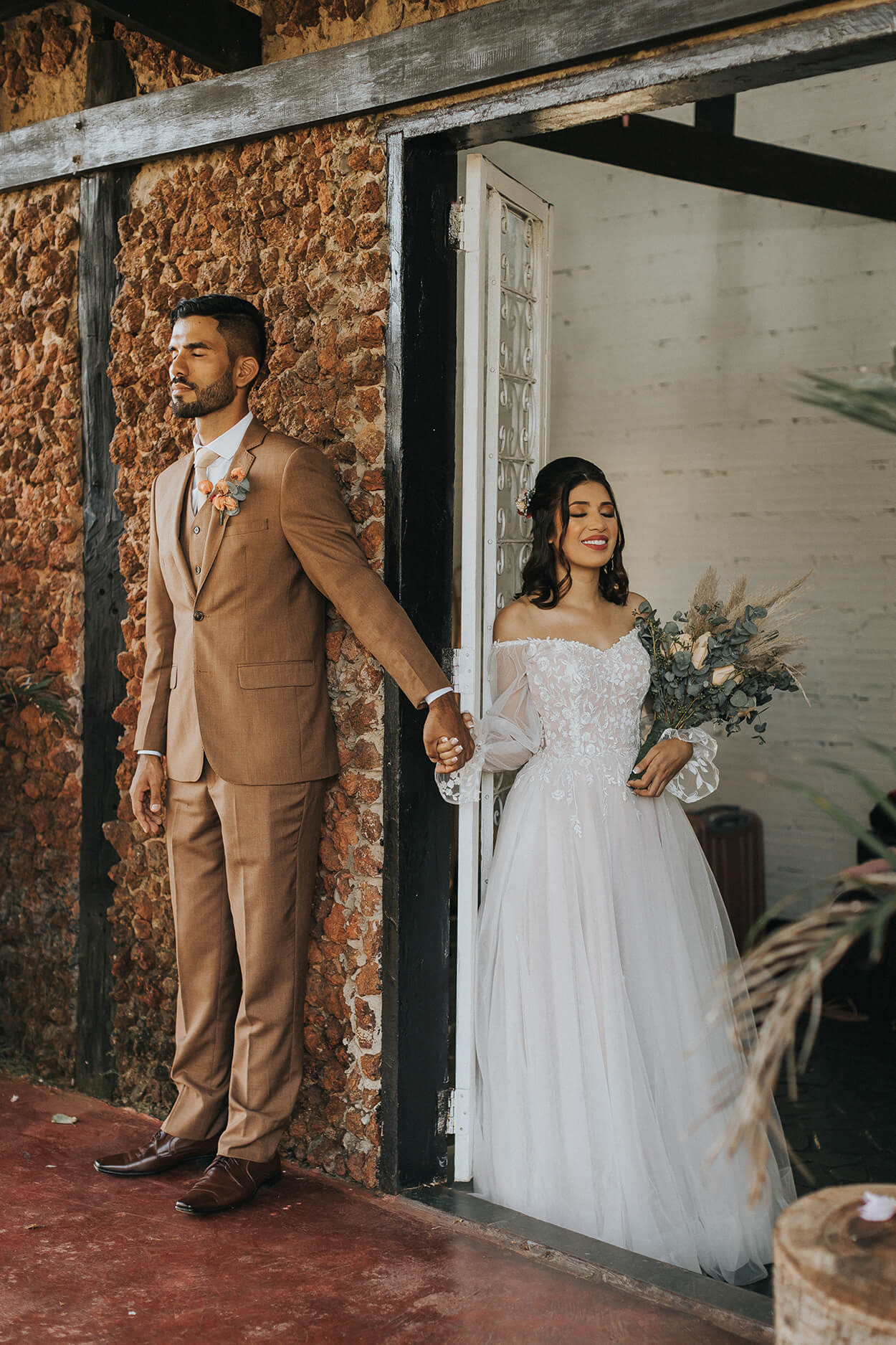 Noivo com terno marrom de mãos dadas com a noiva na porta enquanto os dois estão de olhos fechados