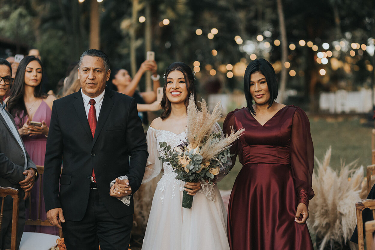Noiva de mãos dadas com o pai de terno preto e gravata vermelha e mãe com vestido bordô acetinado