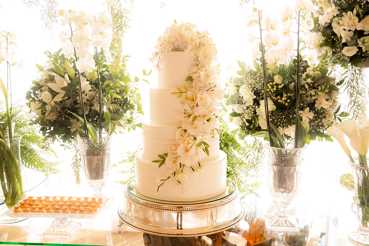 Mesa com bolo de casamento fake com quatro andares e rosas e orquídeas brancas no topo