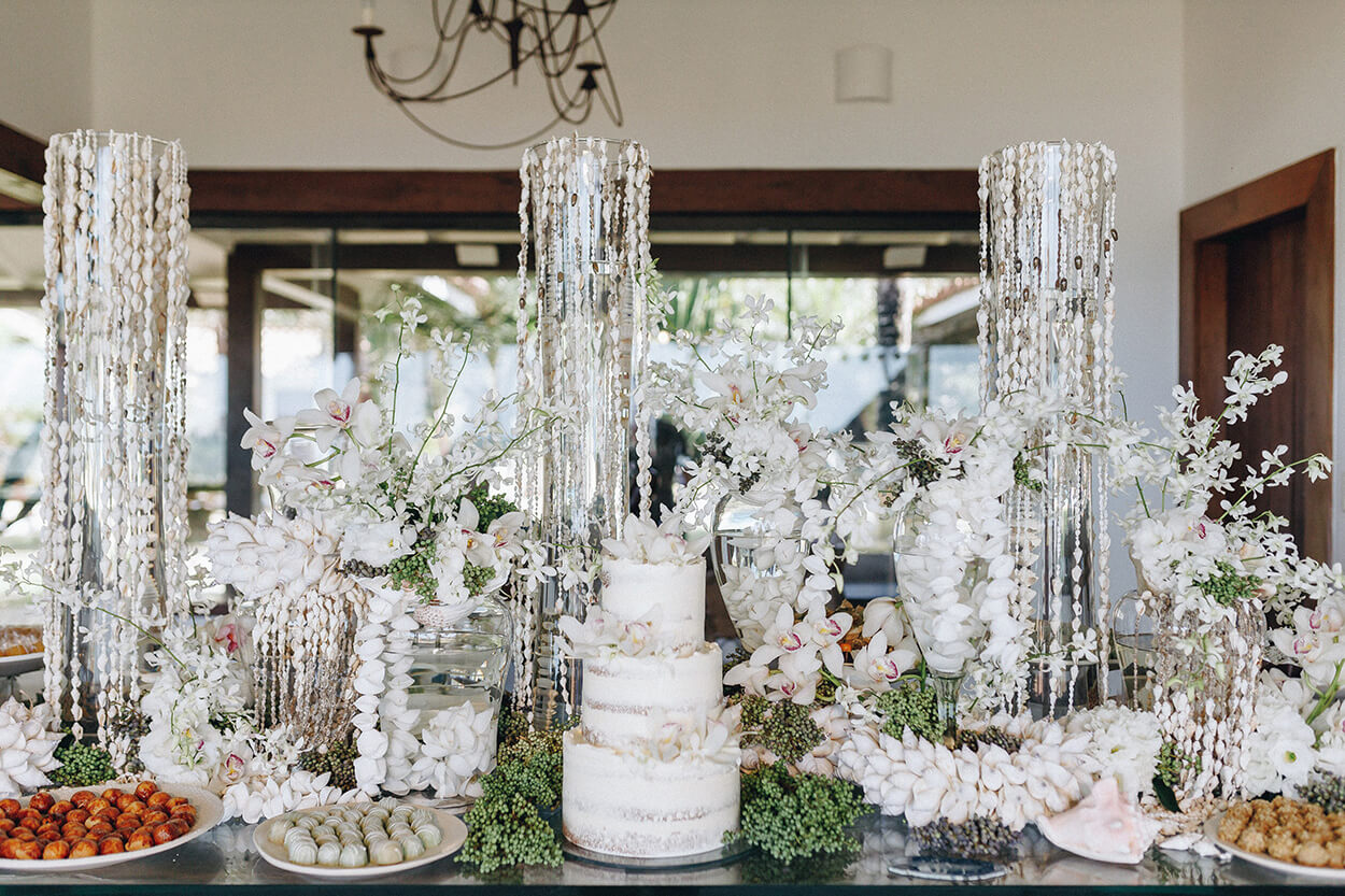 Mesa de vidro com bolo de casamento branco com três andares, pratos com doces de casamento e arranjos brancos