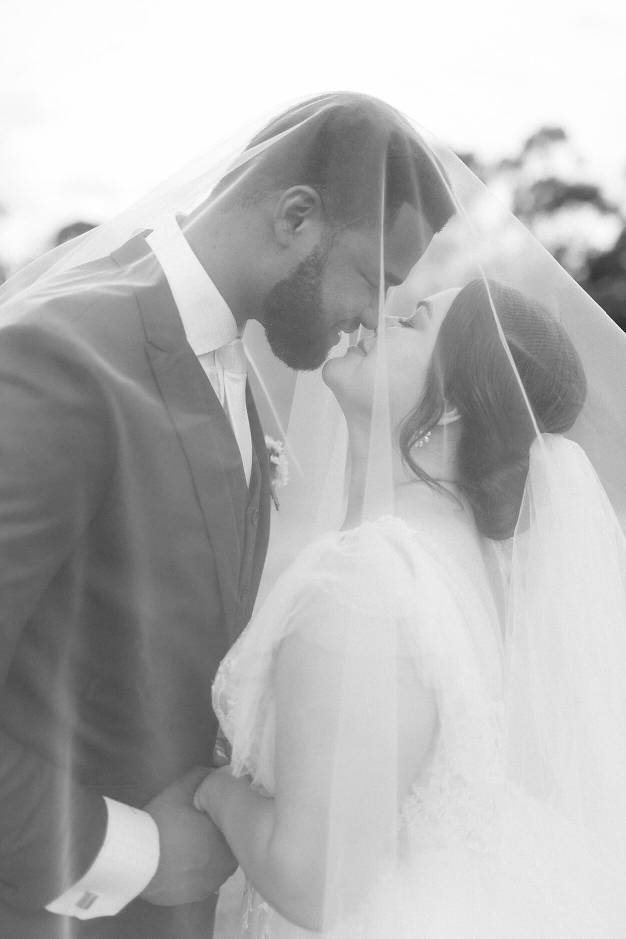Foto em preto e branco dos noivos se beijando debaixo do véu