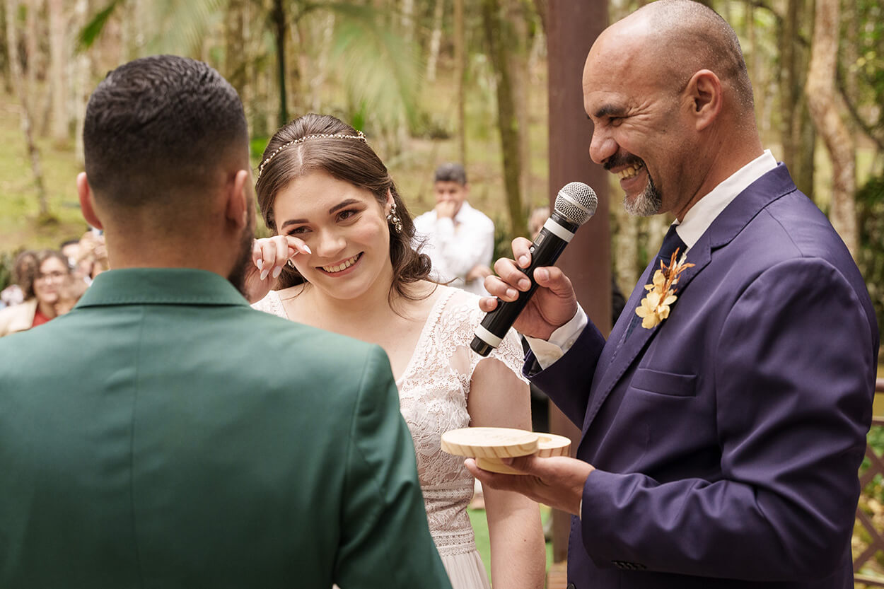 Noiva sorrindo emocionada enquanto celebrante fala ao microfone e segura porta aliança