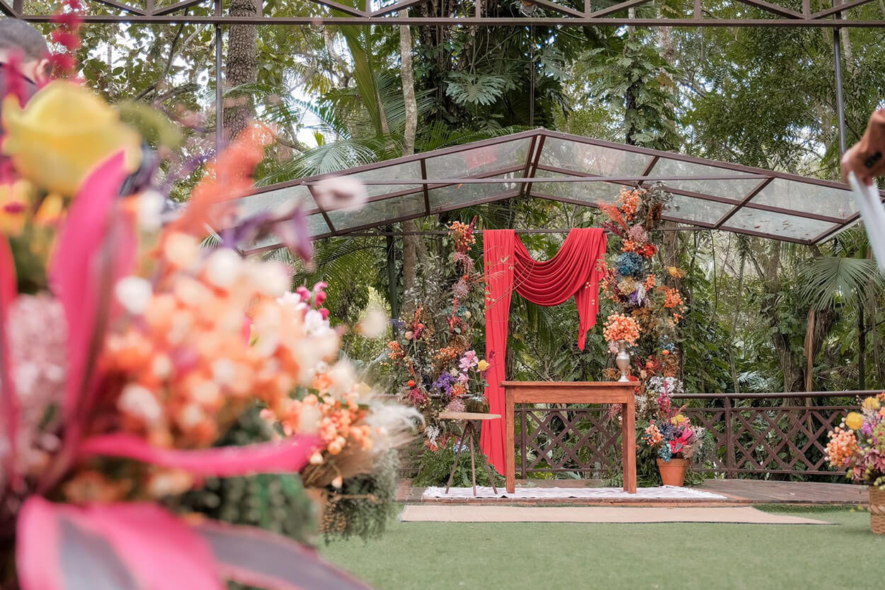 Foto de longe do altar com tecido vermelho pendurado e muitas flores coloridas