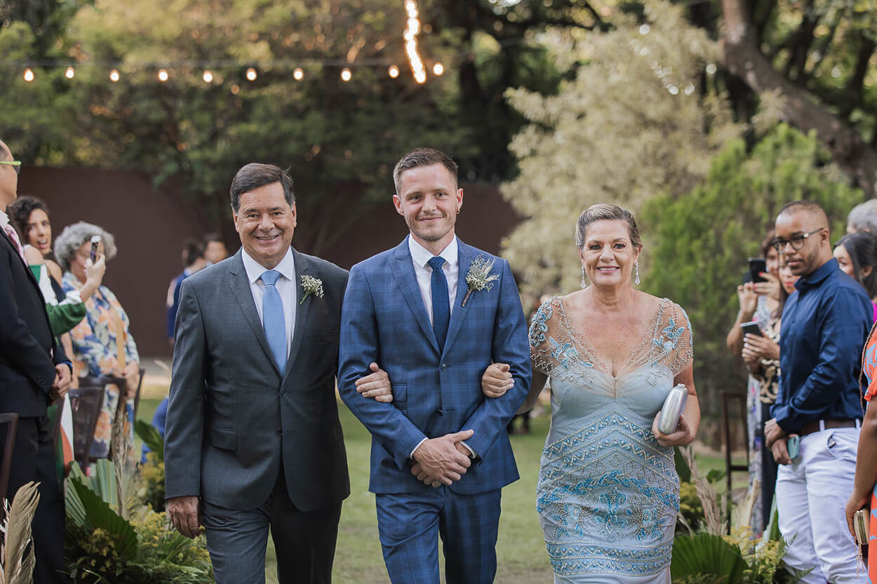 Noivo com terno azul xadrez entrando com o pai de terno cinza e gravata azul serenity e mãe do noivo com vestido azul serenity com bordados e transparência 