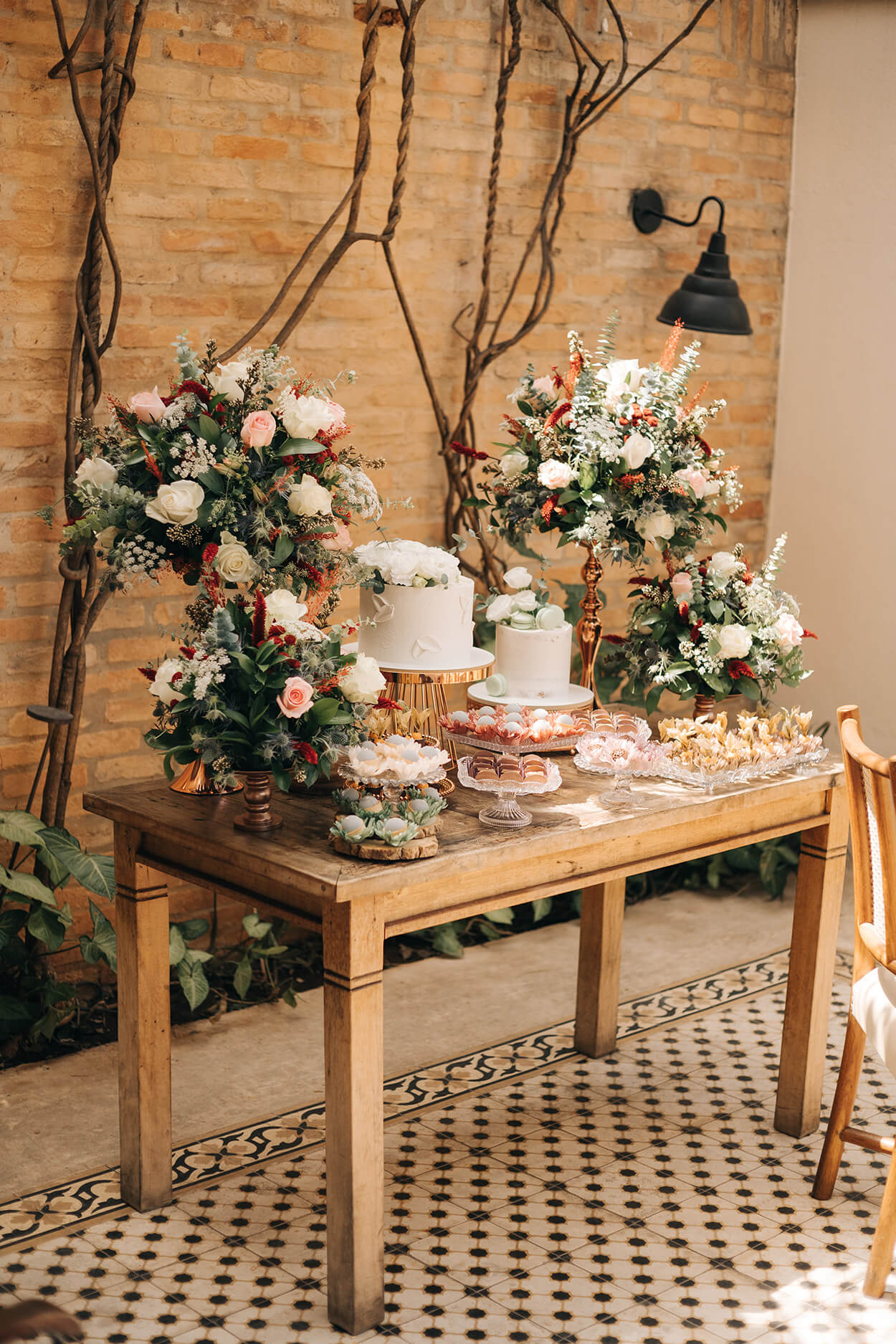 Mesa pequena de madeira com dois bolos redondos de casamento, bandejas com doces de casamento e quatro arranjos com rosas brancas e cor de rosas