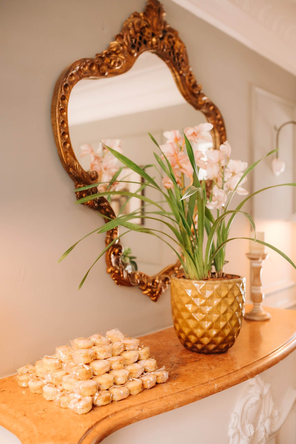 Mesa estreita na parede com bem-casados empilhados e com embalagem transparente. Vaso de flor amarelo ao lado e espelho com moldura antiga na parede