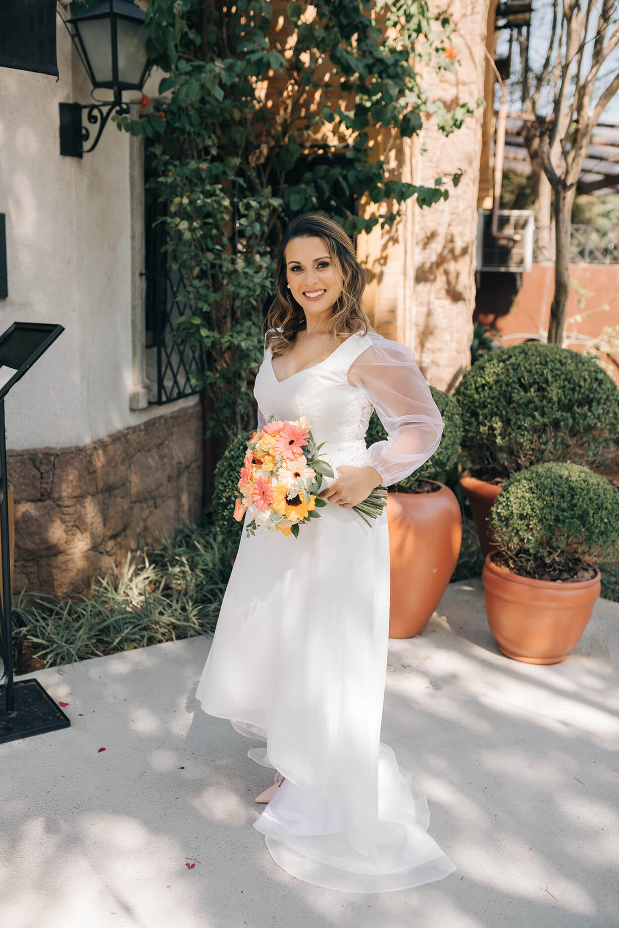 Mulher com vestido de noiva com decote em v e mangas transparentes e bufantes segurando buquê com flores brancas, amarelas e cor de rosa