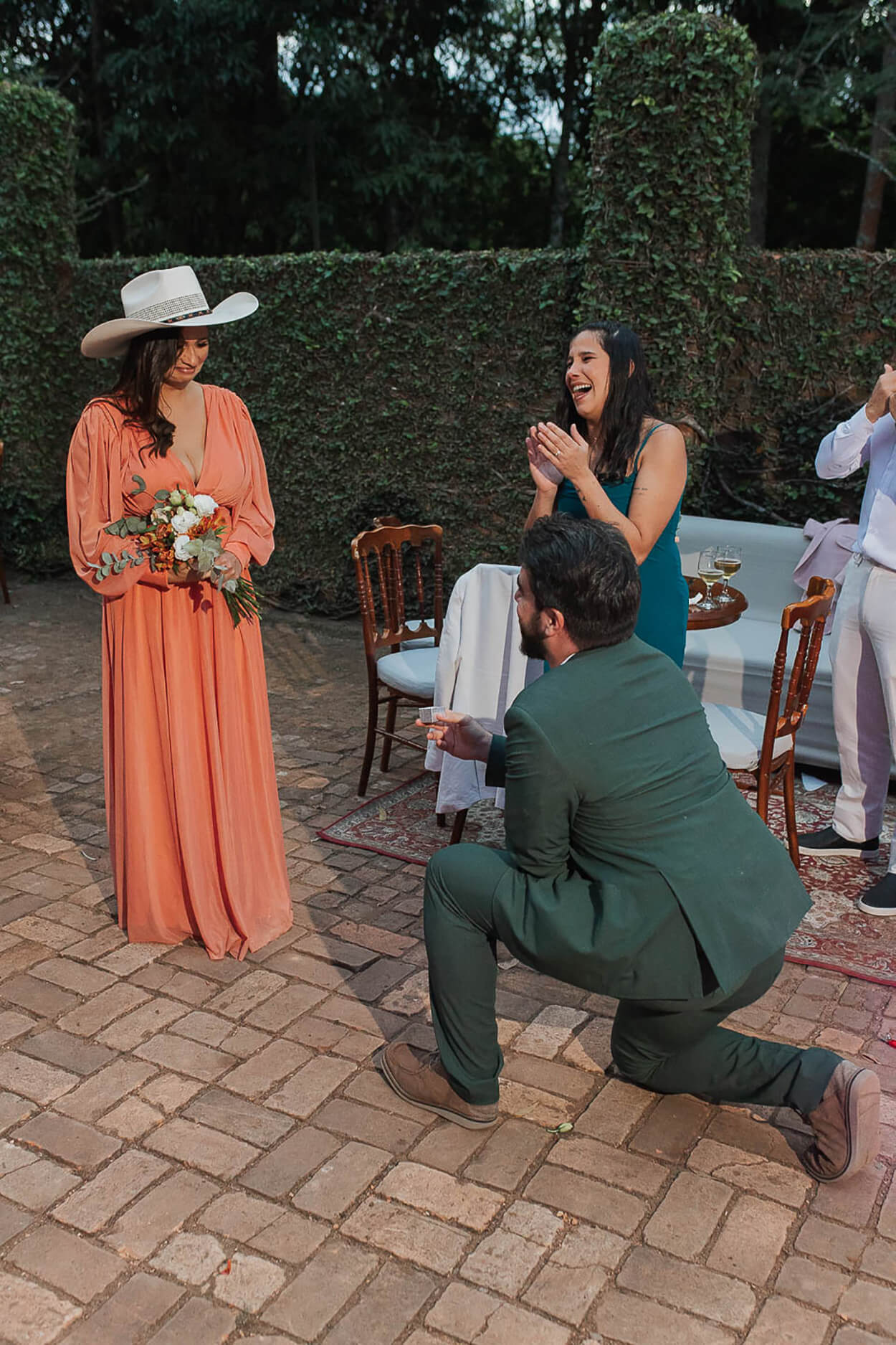 Padrinho com terno verde ajoelhado pedindo madrinhas de vestido laranja em casamento