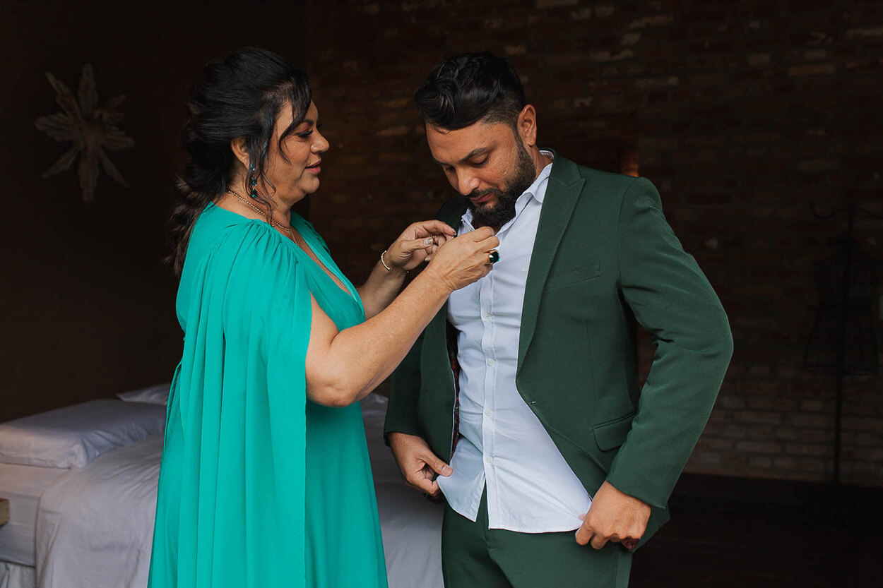Mãe do noivo com vestido turquesa fechando a camisa do filho com terno verde