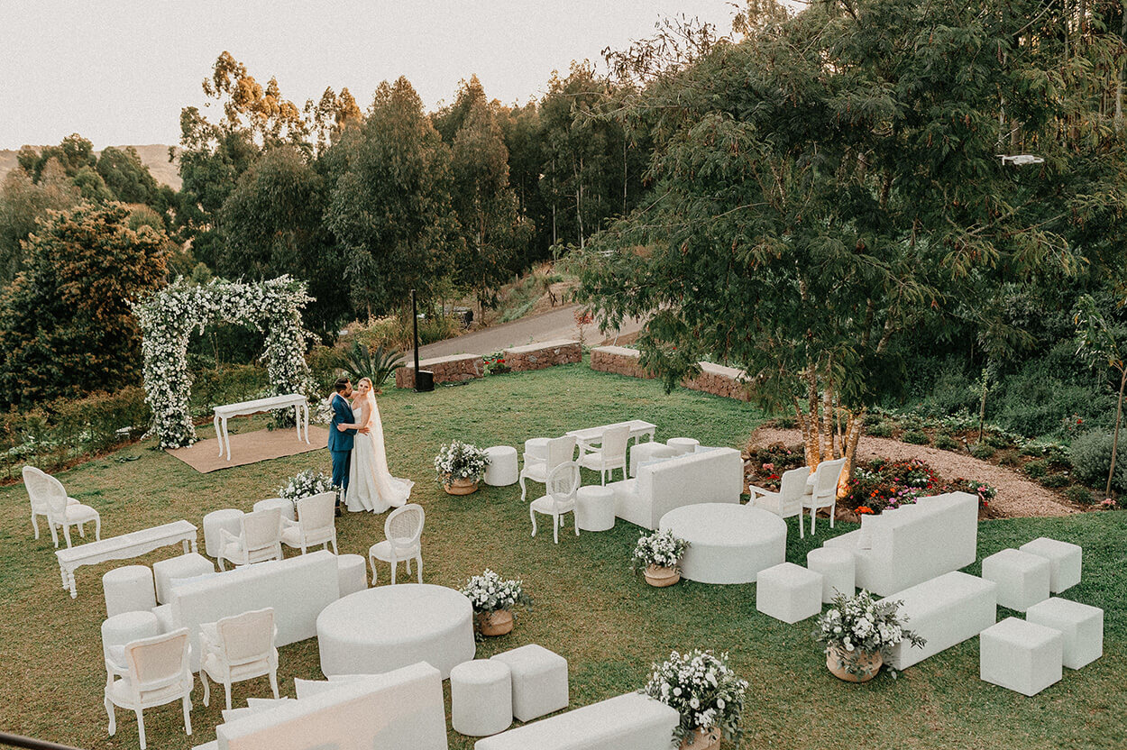 puff, cadeiras brancas e sofás brancos, com e arranjo de flores no campo e noivos no campo
