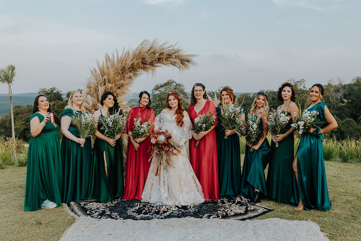 Noiva, mães dos noivos com vestido vermelho longo e madrinhas com vestido verde longo segurando arranjos de flores