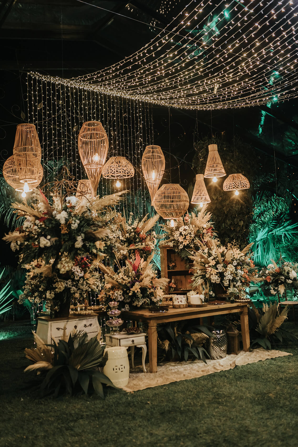 Mesa de madeira e diversos arranjos florais coloridos e luminárias boho acesas e varal de luzes pendente