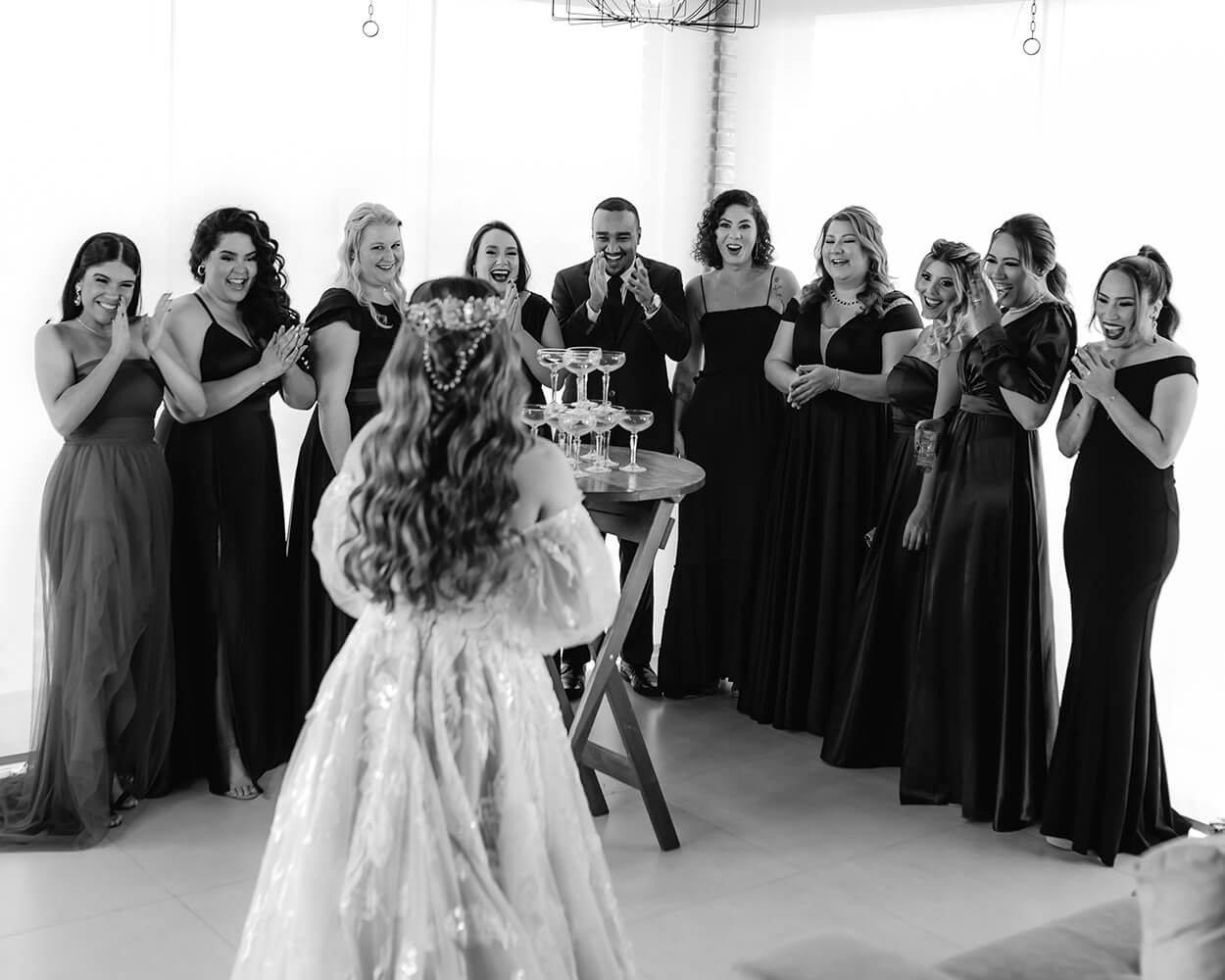 Foto em preto e branco das madrinhas aplaudindo a noiva em uma sala com mesa e torre de drinks