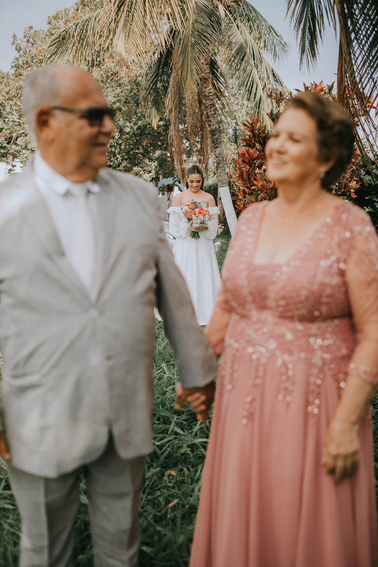 Pai da noiva de terno cinza de mãos dadas com a mãe da noiva usando um vestido rosa brilhante de costas para a noiva