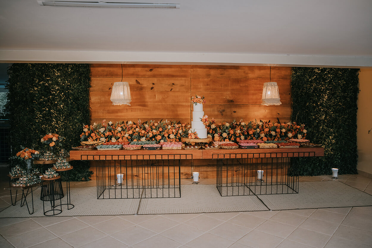 Salão com mesa longa de madeira e metal com bolo de casamento, vários arranjos de flores laranja e bandejas com doces