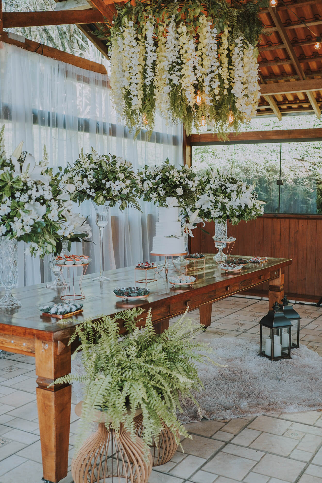 Salão de madeira com mesa com bolo de casamento e earranjos florais brancos