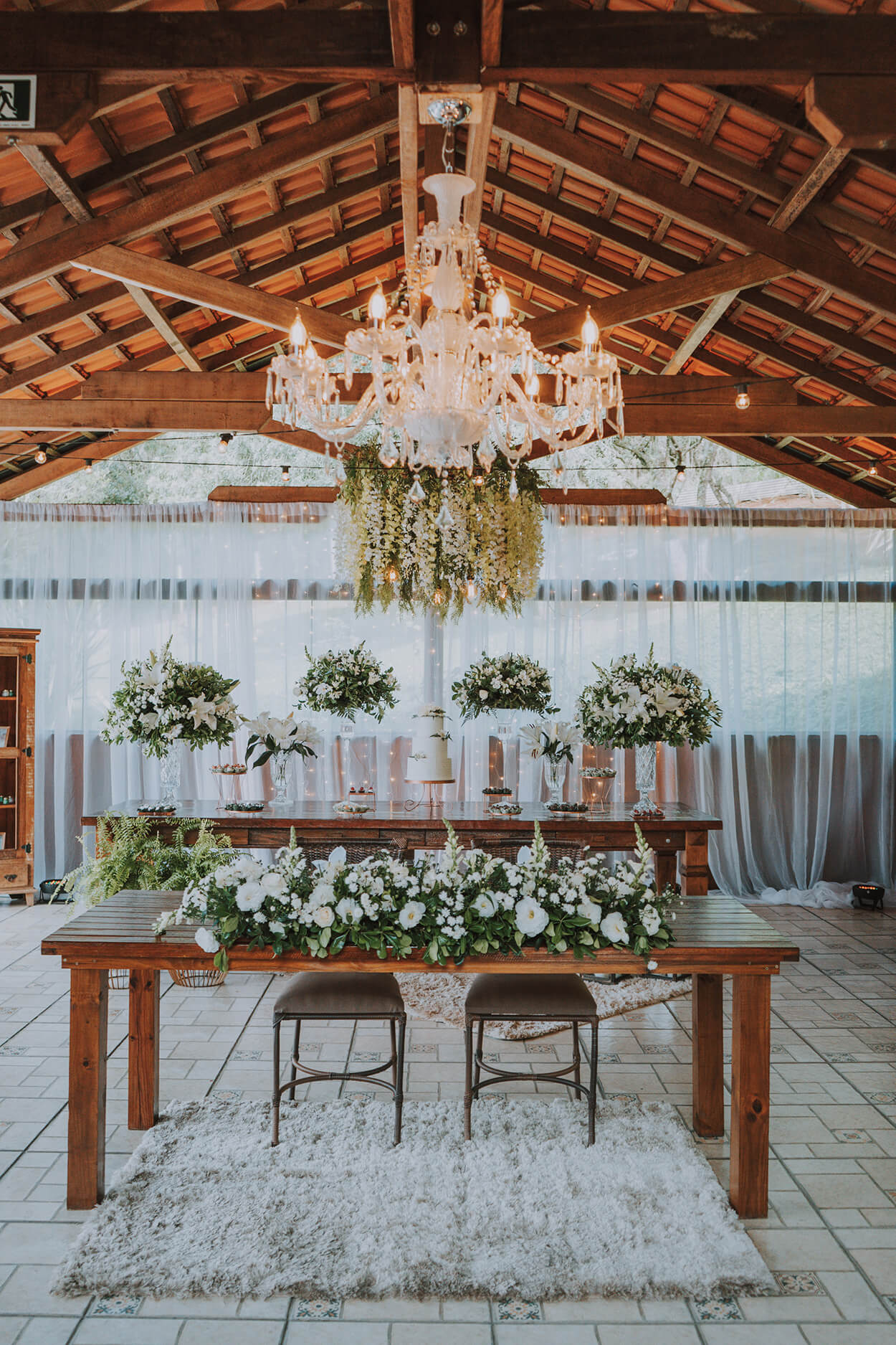 Salão com mesa de madeira com bolo de casamento e arranjos com flores brancas