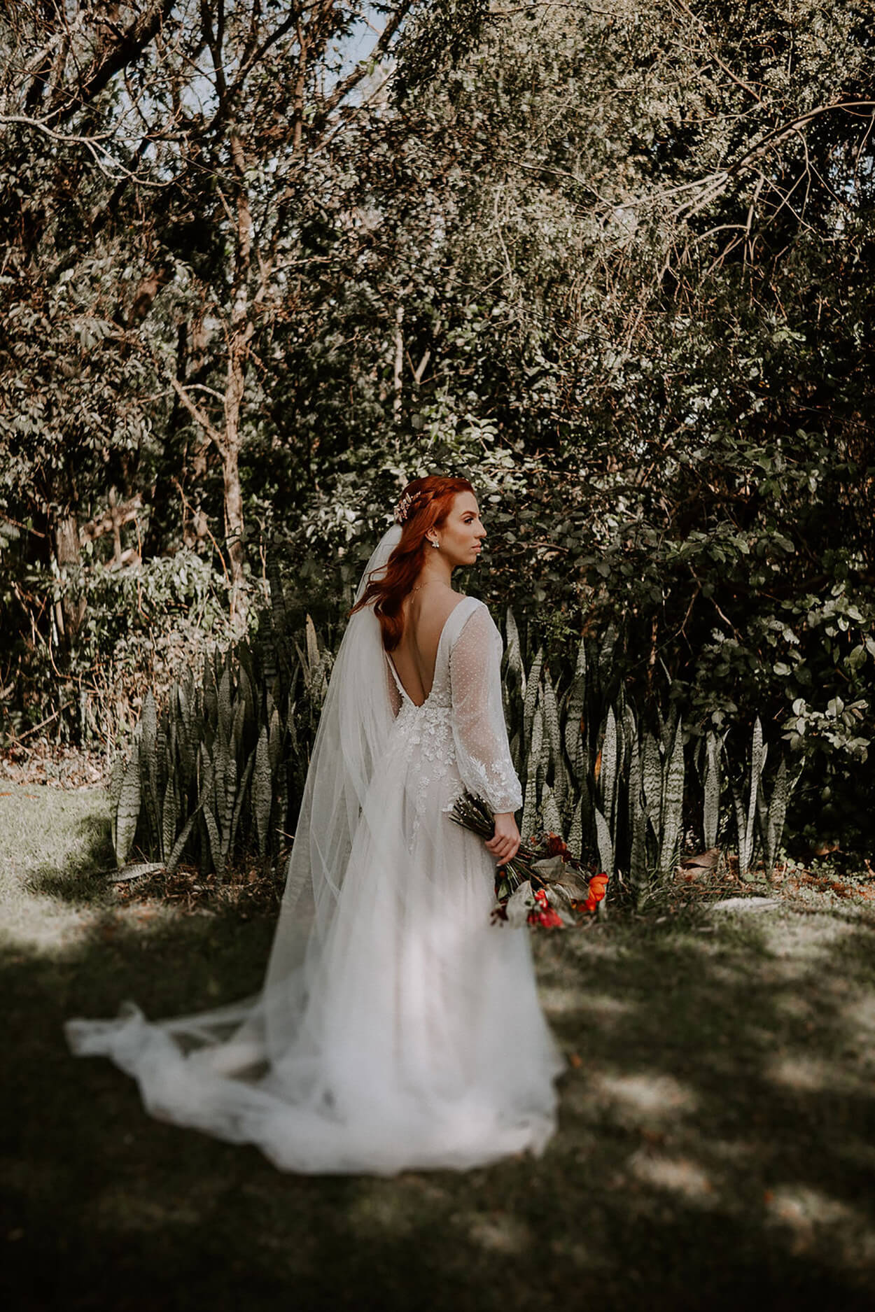 Mulher com vestido de noiva com decote nas costas, manga bufante e véu longo, buquê laranja
