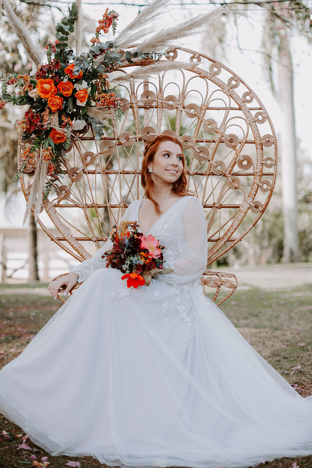Noiva segurando buquê laranja sentada em cadeira pavão estilo boho com flores laranjas e capim dos pampas