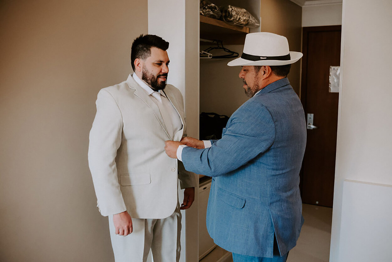 Pai do noivo com terno azul claro e chapéu branco abotoa terno do filho
