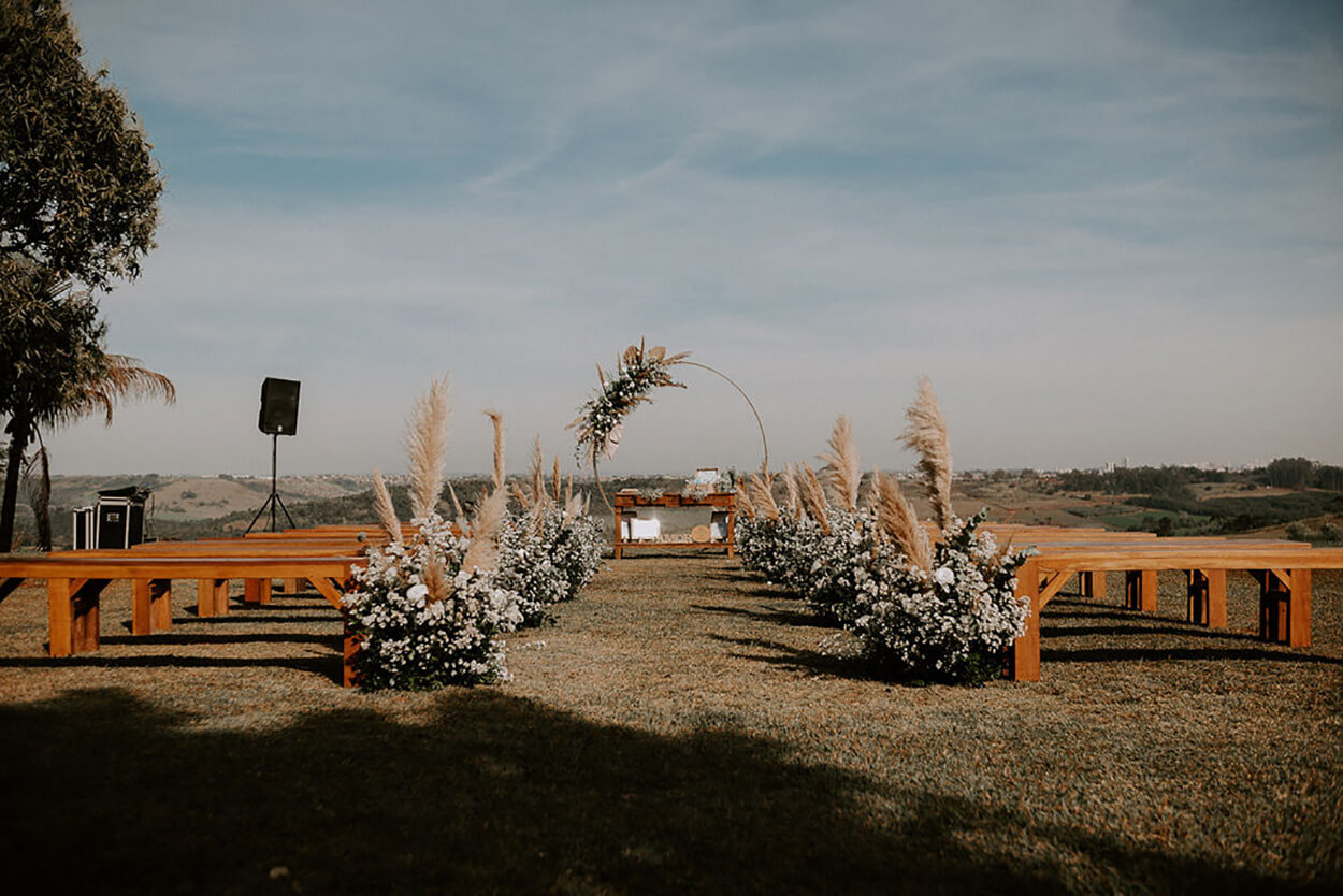 Bancos de madeira com carranjos e flores brancas e capim dos pampas, mesa rústica e altar redondo com capim dos pampas no campo