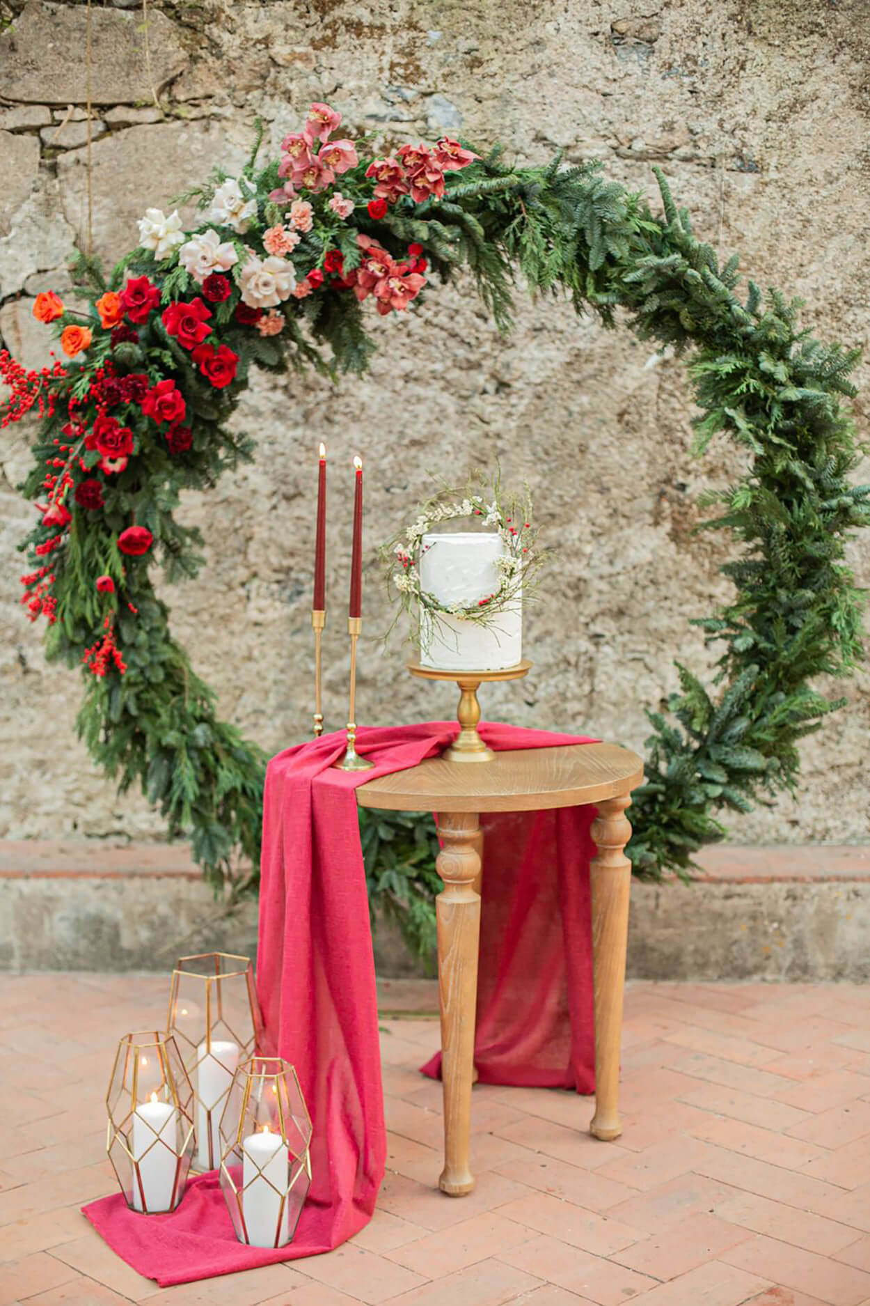 Arco com folhagens e flores vermelhas e brancas com mesa redonda marrom com bolo de casamento e velas